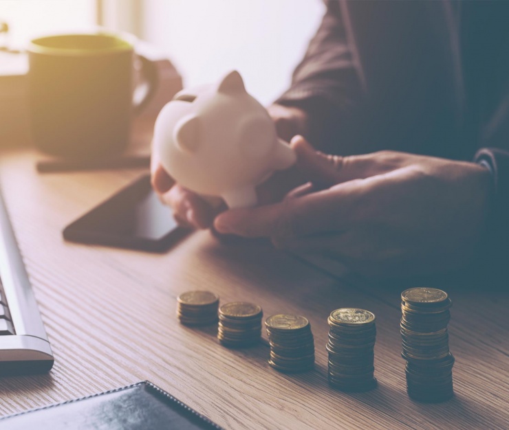 Close-up of a person holding a white piggy bank at a desk, with neatly stacked coins in the foreground representing financial planning and budgeting on a limited income.