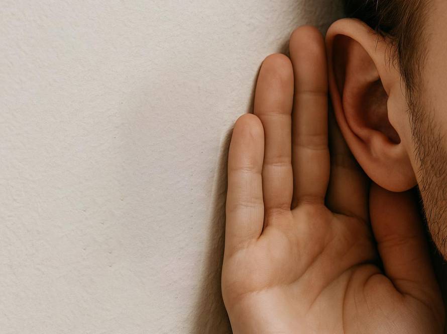 Close-up of a man's ear and hand pressed against a wall, listening intently as if trying to overhear a conversation.